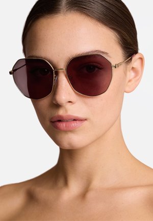 Occhiali da sole ottagonali con lenti rosso scuro e sottili montature in metallo dorato. Design minimalista senza ornamenti visibili.