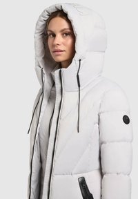 White puffer jacket with a large hood, featuring diagonal quilting, black zippers, and a round logo patch on the left sleeve.