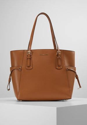 Sac à main en cuir marron avec double poignée, détails de boucles latérales et logo doré subtil sur une surface texturée lisse, posé sur un piédestal blanc.