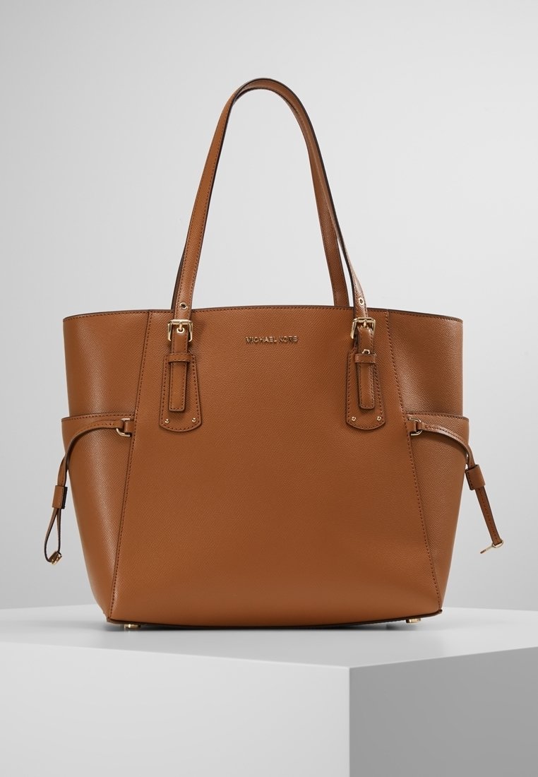 Sac à main en cuir marron avec double poignée, détails de boucles latérales et logo doré subtil sur une surface texturée lisse, posé sur un piédestal blanc.