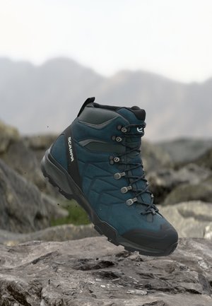 Blue and black hiking boot with rugged sole and laces, positioned above rocky terrain with blurred mountain background.
