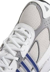 Une chaussure de sport en maille blanche avec des accents gris et bleu, dotée d'un col rembourré, de lacets blancs plats et d'une semelle extérieure texturée pour une meilleure adhérence.