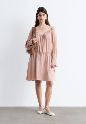 Femme debout vêtue d'une robe rose ample à manches longues, chaussures plates blanches avec bride à la cheville, portant un sac à bandoulière marron sur son épaule gauche.