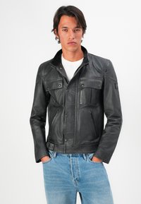 Joven vestido con una chaqueta de cuero negra sobre una camisa blanca, con las manos en los bolsillos de unos jeans azul claro, de pie contra un fondo blanco liso.