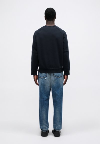 Άνδρας που στέκεται με πλάτη, φορώντας ένα μαύρο φούτερ, μπλε ξεθ�ωριασμένα τζιν με πίσω τσέπες και μαύρα παπούτσια, σε λευκό φόντο.