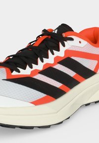 Chaussure de course avec un design blanc, orange et noir, lacets noirs, semelle amortie et semelle crantée pour l’adhérence et le confort.