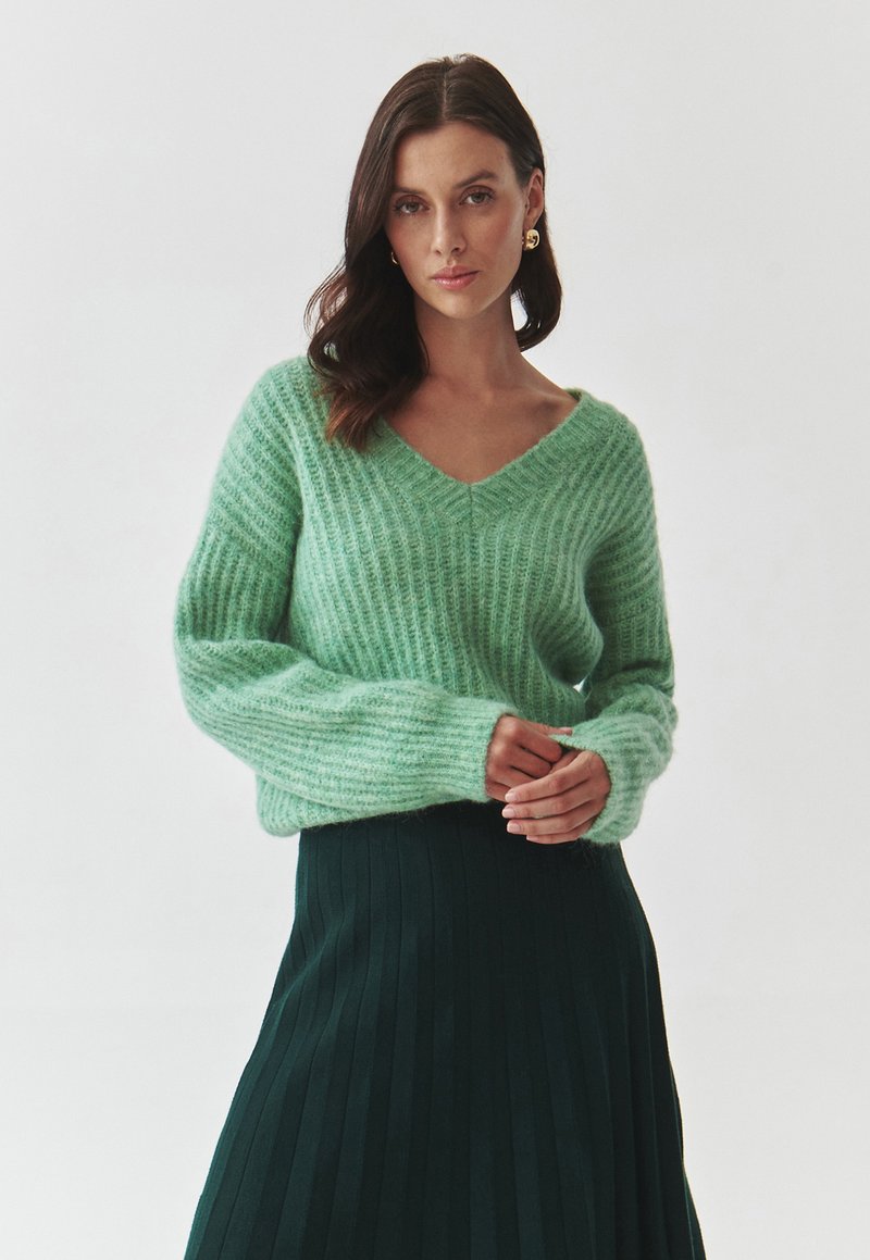 Pull en maille côtelée vert clair avec un col en V et des manches larges, associé à une jupe plissée vert foncé, mettant en valeur la douceur et la texture.