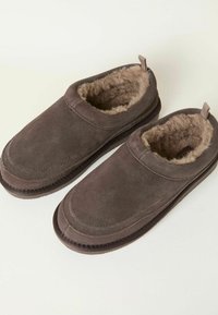 Brown suede slippers with plush lining, slip-on design, rounded toe, and fabric pull tab at the heel. Soft texture and cushioned sole.