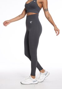 Wysokiej talii ciemnoszare legginsy o gładkiej fakturze z detalem logo w okolicach bioder, w połączeniu z białymi i szarymi sneakersami.