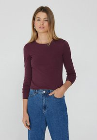 Burgunderfarbene Langarmbluse mit rundem Ausschnitt, glatter Textur, kombiniert mit hochgeschnittenen blauen Denim-Jeans mit Knopfverschluss.