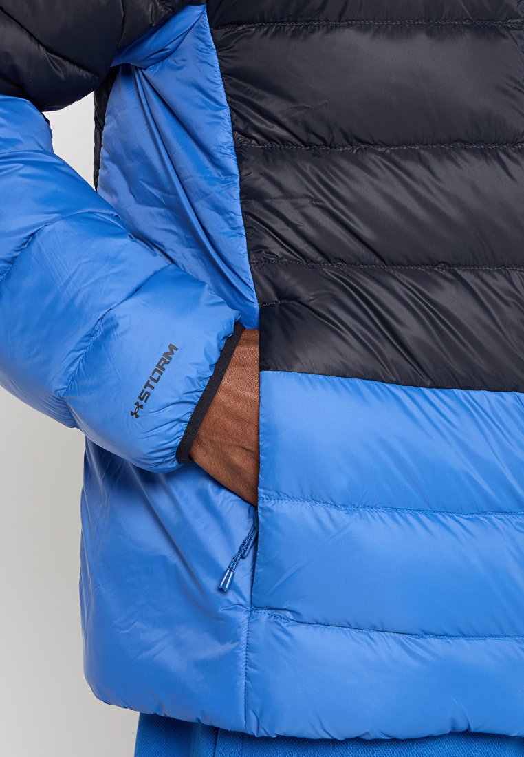 Veste matelassée bleue et noire avec une poche zippée, texture matelassée et un logo sur la manche. Le matériau est brillant et lisse.