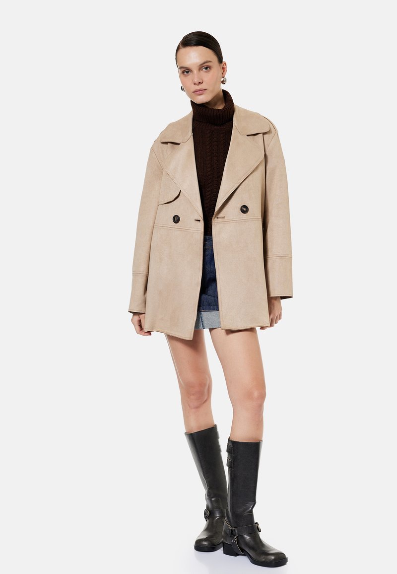 Mujer con abrigo beige cruzado, jersey de cuello alto marrón oscuro, falda vaquera y botas negras hasta la rodilla, de pie sobre fondo blanco.
