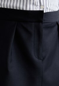 Mørkeblå dressbukser med en glat tekstur, der har en plisseret design, enkelt sidelomme og en flad talje. Knapdetalje øverst.