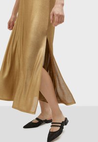 Metalická zlatá maxi šaty s bočními rozparky; hladká textura a splývavý design. Nosené s černými balerínami se špičatou špičkou a třpytivými detaily.