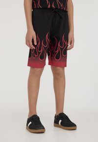 Pantaloni corti neri con motivo a fiamma rossa, in cotone, con vita elastica e cordoncino. Abbinati a sneaker nere con accenti bianchi.