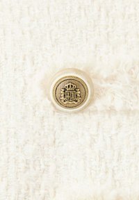 Bouton décoratif doré avec emblème de couronne et de bouclier sur fond de tissu blanc texturé.