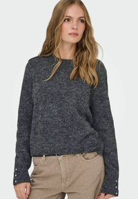 Tamno sivi pleteni pulover s okruglim izrezom i dugim rukavima, s dekorativnim detaljima na manžetama. Uparen s svijetlim bež hlačama.