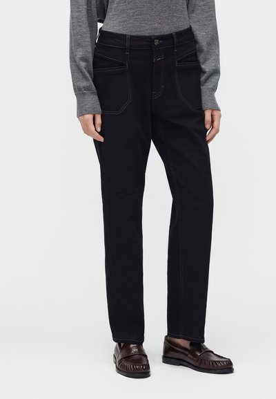 Personne portant un pantalon noir droit avec des poches latérales et avant, un haut gris à manches longues, et des mocassins marron foncé, debout devant un fond uni.