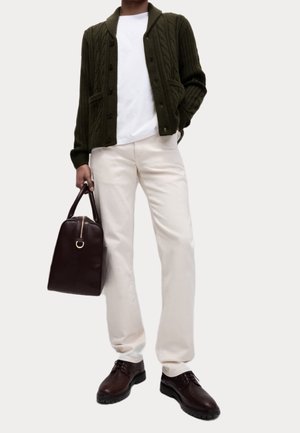 Personne portant un cardigan torsadé vert foncé, une chemise blanche, un pantalon crème, des chaussures en cuir marron, tenant un sac à main en cuir marron dans la main droite.