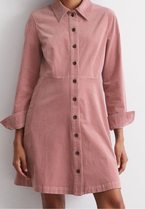 Kvinde iført en støvet lyserød kjole med knapper og lange ærmer, der er rullet op, stående mod en ensfarvet lys baggrund.