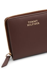 Brown leather wallet with gold hardware, featuring a zipper closure and embossed "TOMMY HILFIGER" logo on the front. Compact rectangular design.