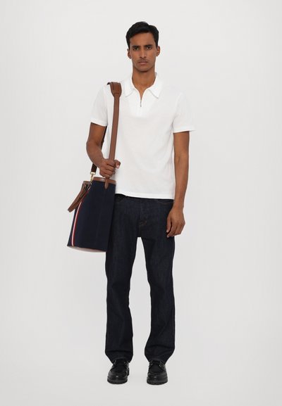 Balta polo marškinėliai, tamsios džinsinės kelnės, laikanti tamsiai mėlyną krepšį su rudos odos dirželiu ir raudonai baltomis dryžuotėmis. Juodi batai.