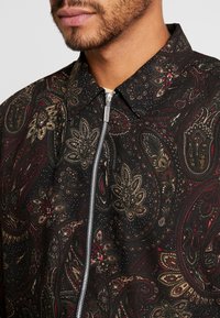 Man in een donkere ritsblouse met een ingewikkeld paisleypatroon in rood, beige en zwart, en een klassieke kraag.