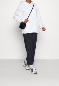 Bílá oversized mikina s kapsou na přední straně a stahovací kapucí; spárovaná s námořnickými kalhotami s pruhy a černým křížovým batohem. Na nohou sportovní obuv.