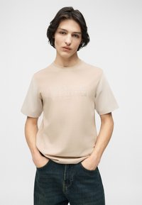 Νέος άνδρας με σκούρα μαλλιά που φοράει μπεζ T-shirt Jack & Jones Premium και σκούρο τζιν, με τα χέρια στις τσέπες, ουδέτερο φόντο.