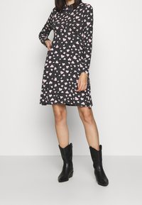 Svart klänning med långa ärmar, prydd med ett mönster av rosa hjärtan och prickar. A-linjens snitt har fickor. Matchas med svarta ankle boots.