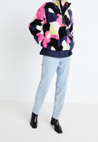 Farverig fleecejakke med abstrakte former i pink, navy og grøn, med en sort lynlåns-krave. Sammen med lyseblå jeans og sorte støvler.