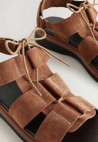 Sandalias de ante marrón con un diseño de tiras, que presentan lazos en nudos, suelas planas y acentos rectangulares distintos. Plantillas y detalles de costura en negro.