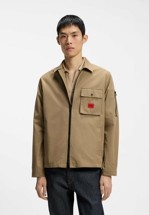 Junger Mann trägt eine beige Jacke mit Reißverschluss, Brusttasche und rotem Logo-Patch, kombiniert mit dunkelblauen Jeans, steht vor einem einfarbigen Hintergrund.