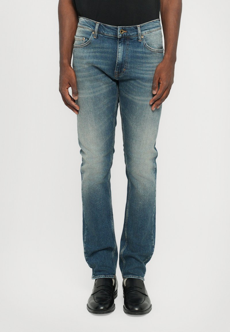 Jeans iz denima v gradientnem modrem pranju, s ravnim krojem nogavic, petimi žepi in subtilnim zbledelostjo. Črne čevlje dopolnjujejo videz.