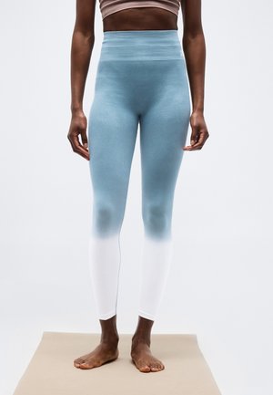 Persona de pie descalza sobre una estera, vistiendo leggings de cintura alta con degradado azul a blanco y un top corto beige.