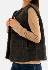 Gilet in pile grigio scuro con finitura texturizzata, design senza maniche e tasche laterali, indossato sopra una maglietta beige chiaro a maniche lunghe e jeans.