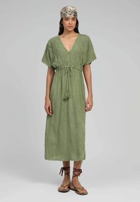 Robe en lin vert olive avec des manches larges, un décolleté en V profond et une taille avec cordon de serrage. Assortie à des sandales à lanières marron et un bandeau à motifs.