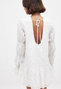 Balta suknelė su gėlėmis siuvinėjimu, ilgomis pūstomis rankovėmis ir gilia nugaros iškirpte su raišteliu. Platėjanti apatinė dalis suteikia žaismingą siluetą.