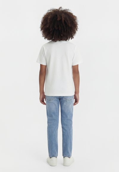 Bērns ar cirtainiem matiem stāv ar muguru, valkājot vienkāršu baltu īsroku T-kreklu, gaiši zilas džinsas un baltas sporta kurpes.