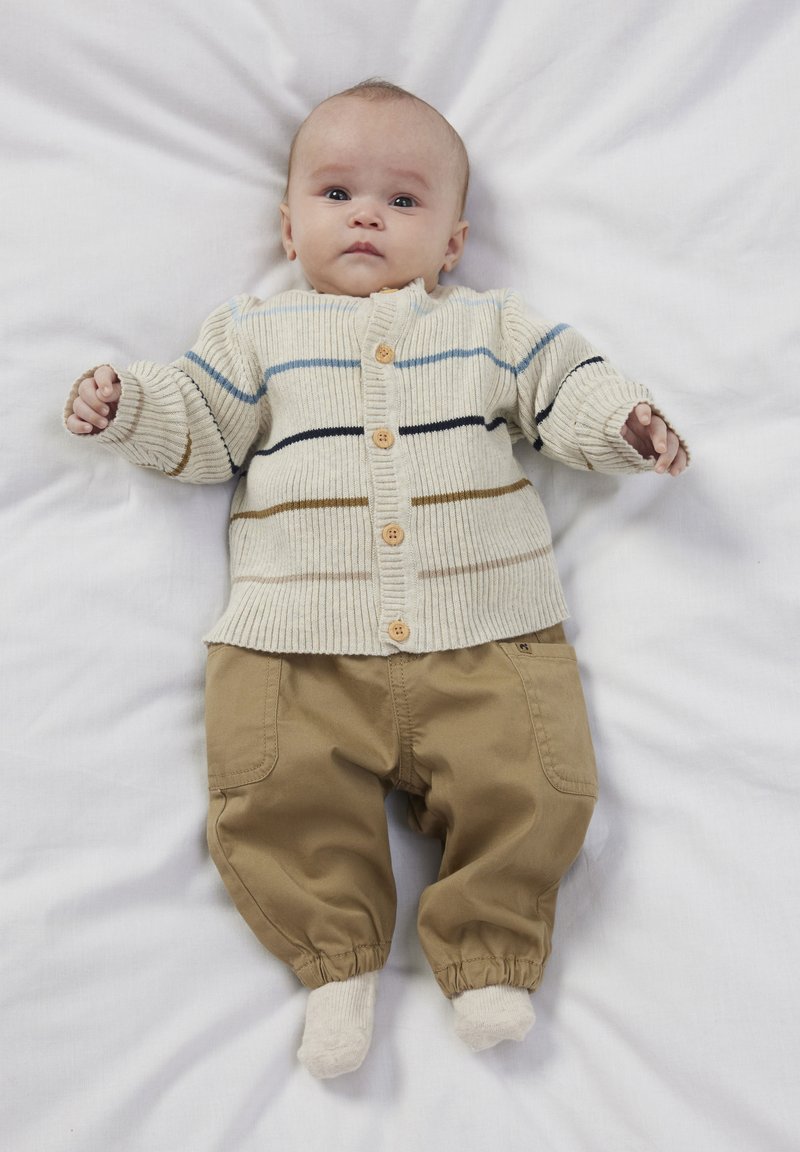 Pleteni krem kardigan z modrimi in rjavimi črtami, z lesenimi gumbi, v kombinaciji s svetlo rjavimi bombažnimi hlačami in svetlimi nogavicami, razporejen na belem ozadju.