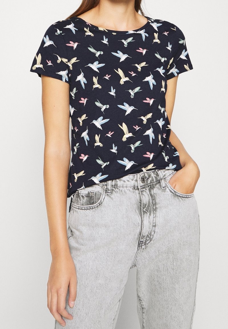Femme portant une chemise bleu marine à manches courtes avec des motifs d'oiseaux aux couleurs pastel et un jean taille haute gris clair, une main dans la poche.