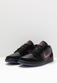 Baskets noires en cuir et daim, avec un Swoosh Nike rouge et des détails texturés. La semelle est de couleur bleue translucide.