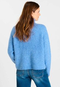 Vrouw met lang bruin haar, gekleed in een lichtblauwe harige trui en een blauwe spijkerbroek, staande met haar gezicht naar rechts tegen een witte achtergrond.