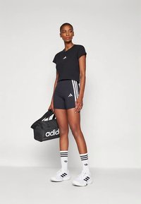 T-shirt preta de manga curta e calções de cintura alta com três riscas brancas; combinado com meias e sapatos brancos, e uma bolsa de desporto preta.