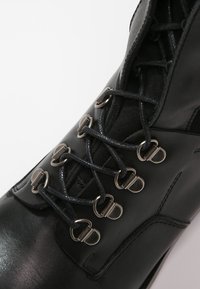 Bota de cuero negra con un acabado liso, ojales metálicos y cordones negros. Incluye una punta redondeada y detalles de costura sutiles.