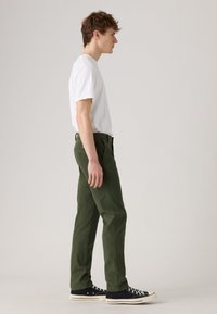 Young man standing sideways wearing white t-shirt, olive green pants, and black low-top sneakers on plain light background.