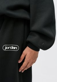 Χέρι που ξεκουράζεται δίπλα σε μαύρες παντελόνες Jordan με άσπρο λογότυπο και ταιριαστή μαύρη μανσέ τα με ανάγλυφο λογότυπο Jumpman στον γάντζο.