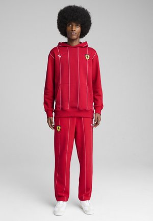 Hoodie rouge avec rayures blanches, poche kangourou et logo Ferrari ; associé à un pantalon de survêtement rouge assorti présentant le logo et des rayures blanches.