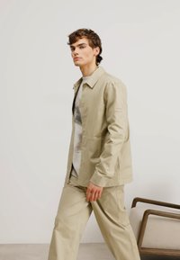 Chaqueta de botonadura ligera color beige claro con mangas largas, combinada con pantalones a juego. La tela parece suave, con un diseño minimalista.