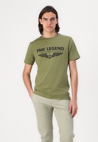 Olivegrüne Baumwoll-T-shirt mit schwarzem Aufdruck "PME LEGEND AMERICAN CLASSIC" und Flügel-Design. Kurze Ärmel und Rundhalsausschnitt.